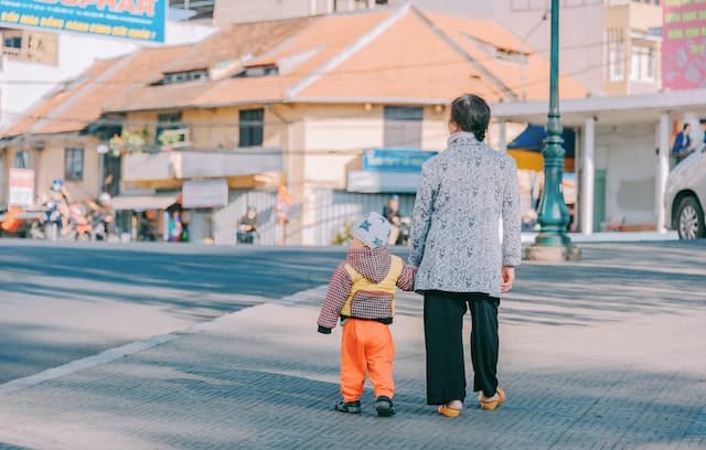 An older woman in a light blue jacket walks along a street holding the hand of a toddler in orange pants.