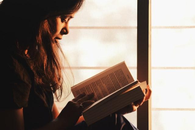 Young woman reading a book illuminated by light shining through a window.